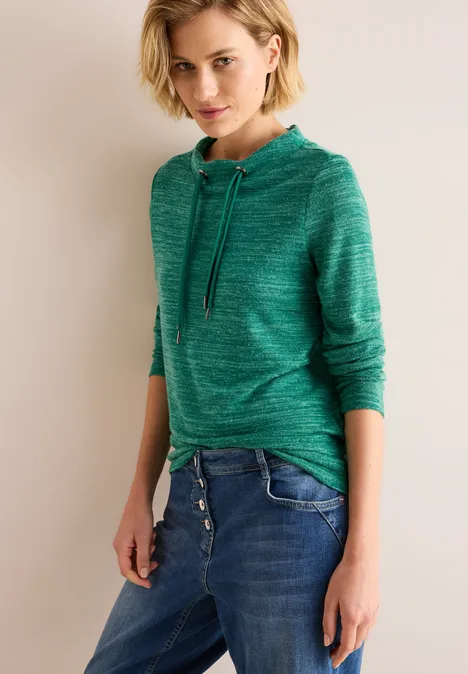 Frau mit grünem Pullover und blauer Jeans steht vor hellem Hintergrund.